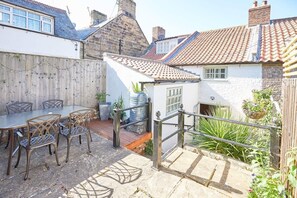 A patio with a table and chairs, a small white building with a tiled roof, and a wooden fence.