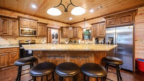 A wooden cabin-style kitchen with a large island, bar stools, stainless steel appliances, and wooden cabinets. - Luxury Broken Bow Lodge | 7 King Suites | Sleeps 20+ (Broken Bow)