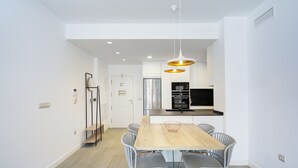 A modern kitchen with a stainless steel refrigerator, a built-in oven, and a black microwave. There is a wooden dining table with four chairs and a hanging light fixture above it.