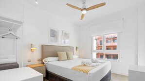 A modern bedroom with a bed, bedside tables, a ceiling fan, and a window with a view of buildings.