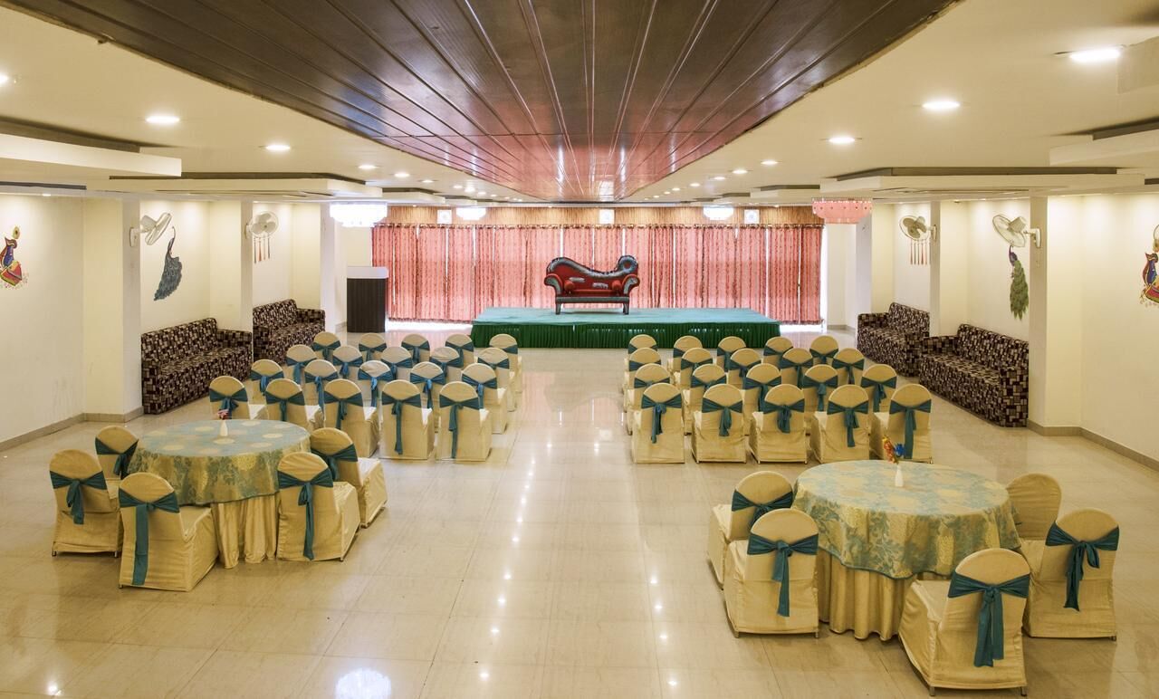 A banquet hall with round tables and chairs, a stage with a red backdrop, and wall-mounted fans.