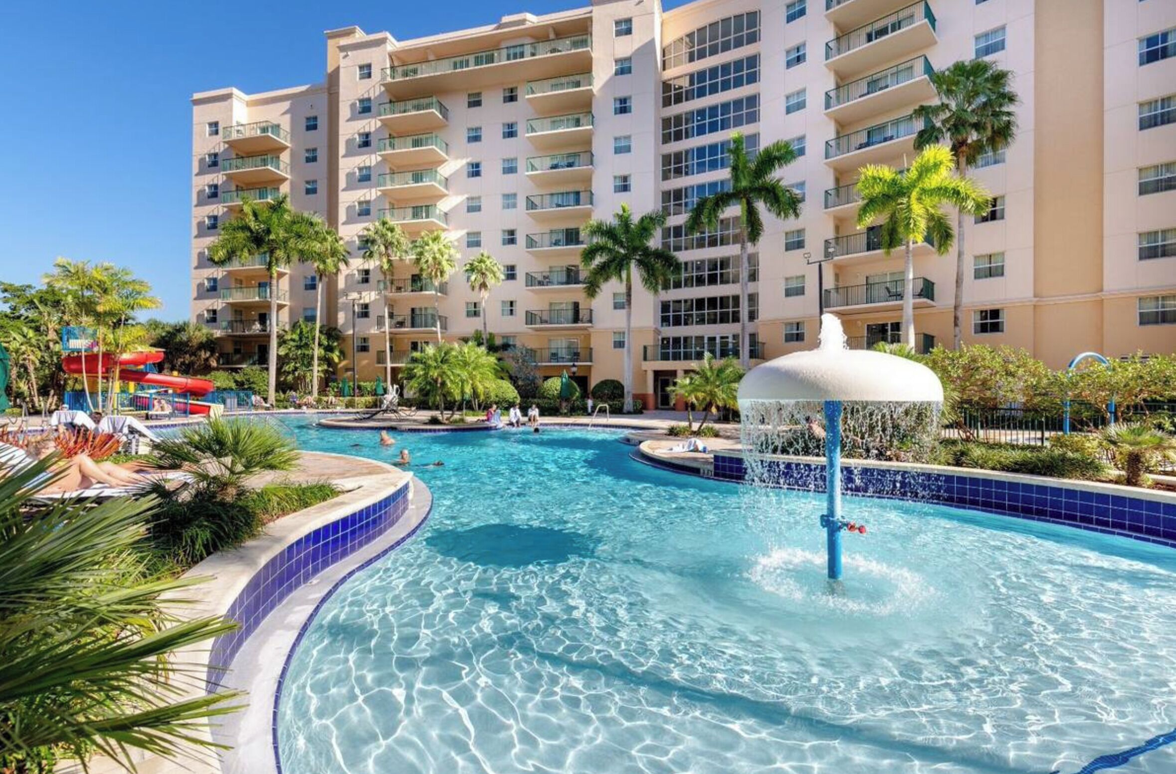 A hotel with a large swimming pool, palm trees, and a water fountain.