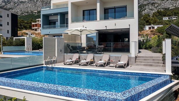 A modern two-story building with a swimming pool, outdoor seating, and a view of rocky mountains.