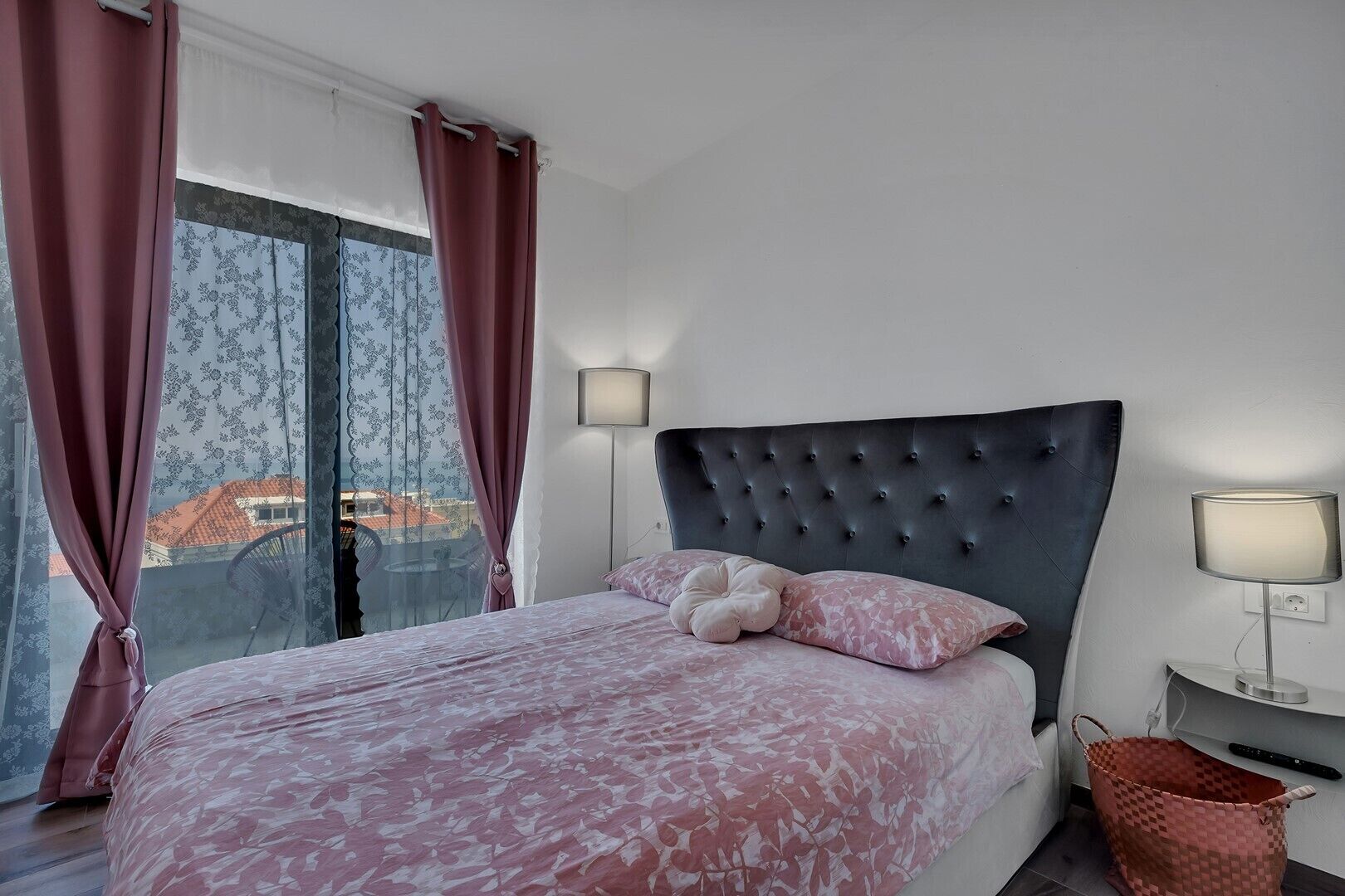 A bedroom with a large bed, two bedside lamps, a window with curtains, and a view of buildings outside.