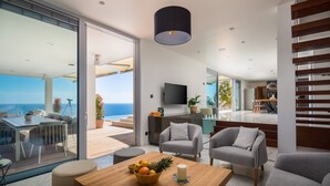 Ein modernes Wohnzimmer mit einem großen Glasfenster, das auf einen Balkon mit Meerblick führt. Es gibt einen Holzesstisch mit Stühlen, ein Sofa und einen an der Wand montierten Fernseher.
