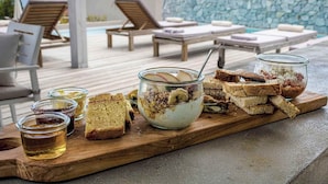 A breakfast buffet with bread, yogurt, and honey on a wooden board, overlooking a pool area with lounge chairs and a stone wall.