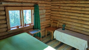 A cabin with a bed, a small table, a chair, a window with curtains, and a view of the forest. - Lomakoti kauniilla näkymillä Saimaan rannalla (Savonlinna)