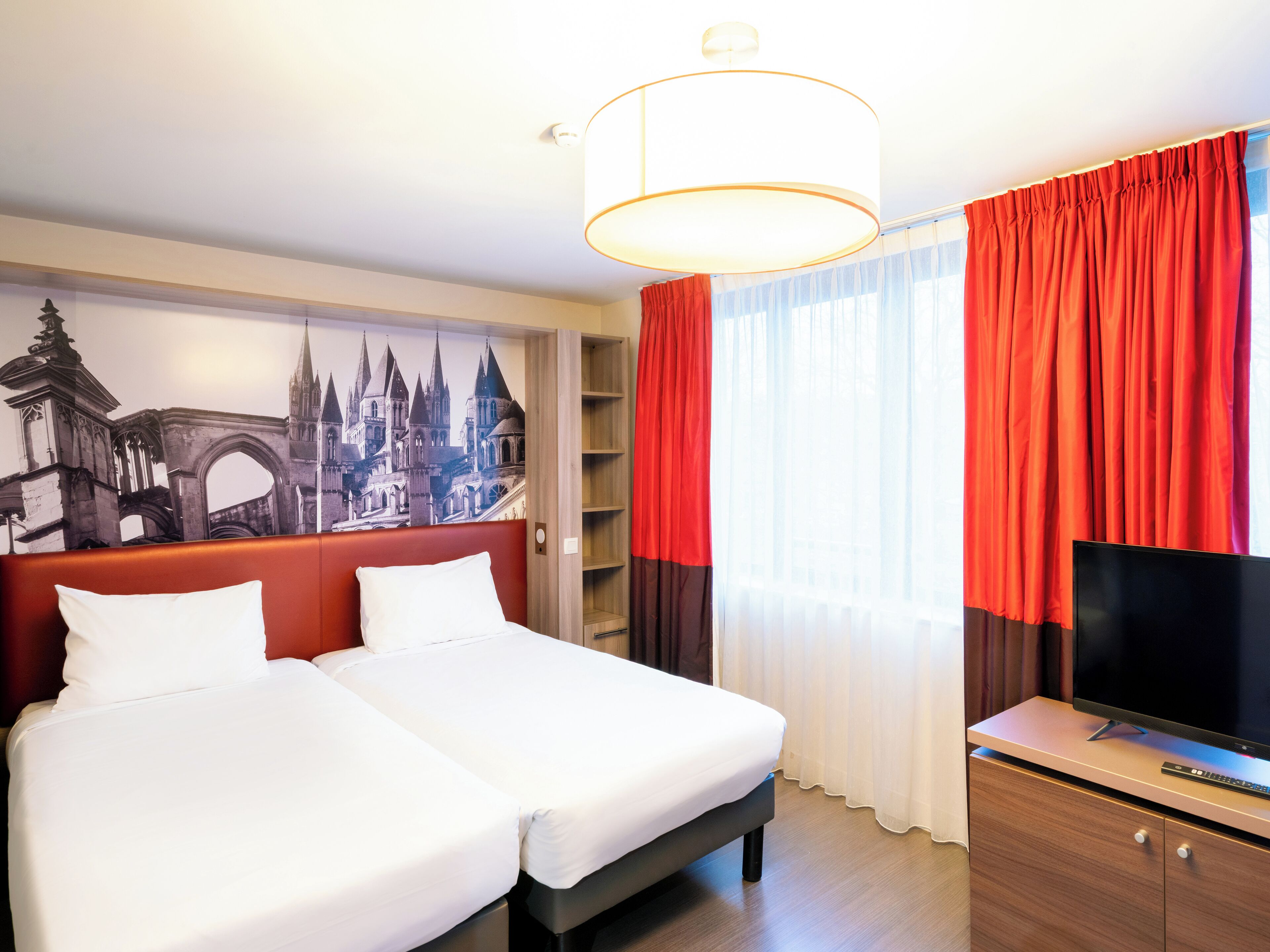 A hotel room with two beds, a TV, a large window with red curtains, and a mural of a cityscape.