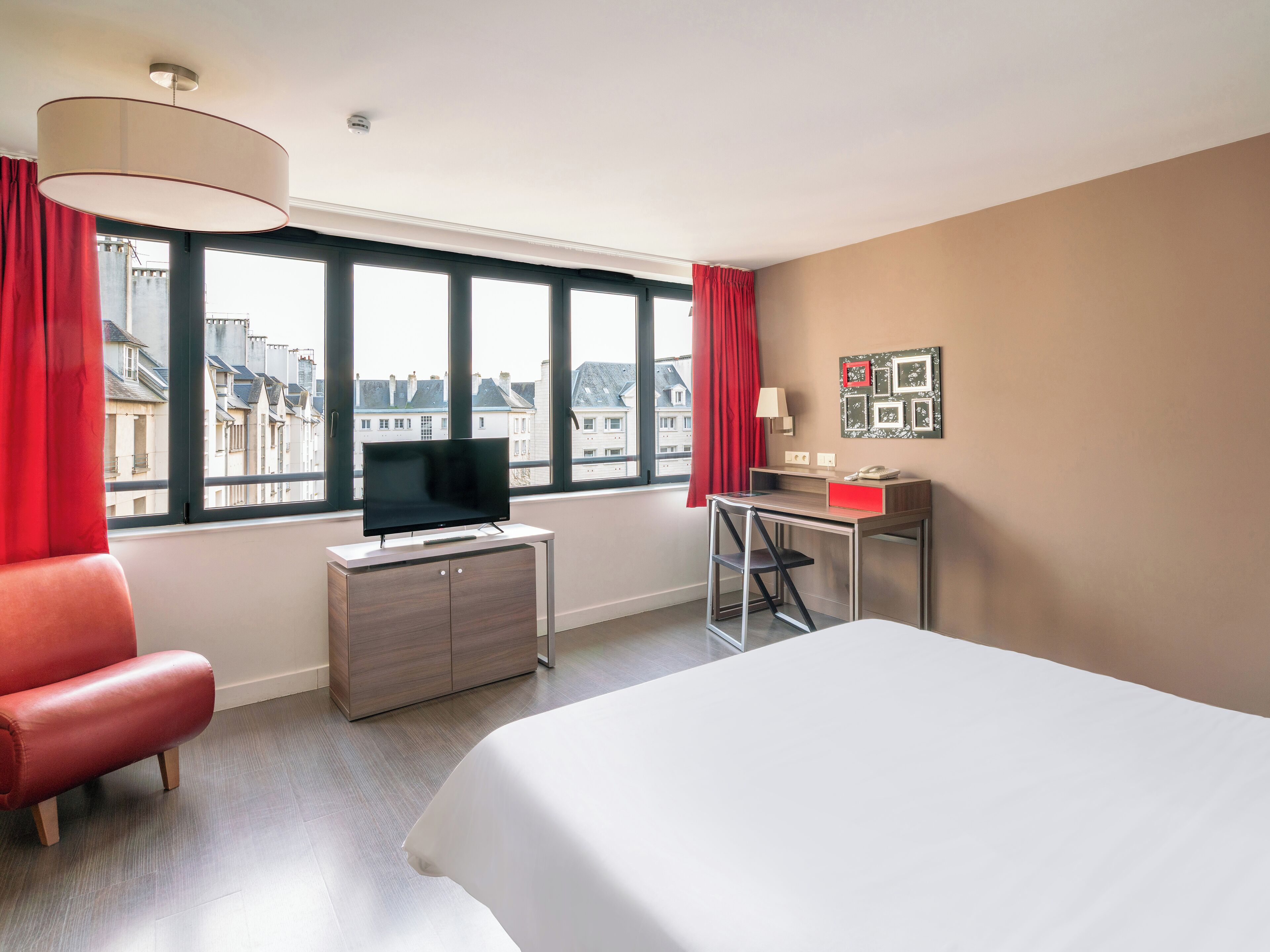 Ein Hotelzimmer mit einem Bett, einem Schreibtisch mit Computer, einem Fernseher auf einer Kommode und einem Fenster mit Blick auf Gebäude.