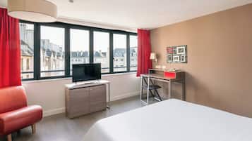 Ein Hotelzimmer mit einem Bett, einem Schreibtisch mit Computer, einem Fernseher auf einer Kommode und einem Fenster mit Blick auf Gebäude.