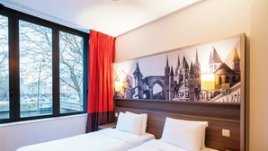 A hotel room with two beds, a large window with a view of a cityscape, and a wall-mounted painting of a historical building. - B&B HOME Caen Centre Gare (Caen)