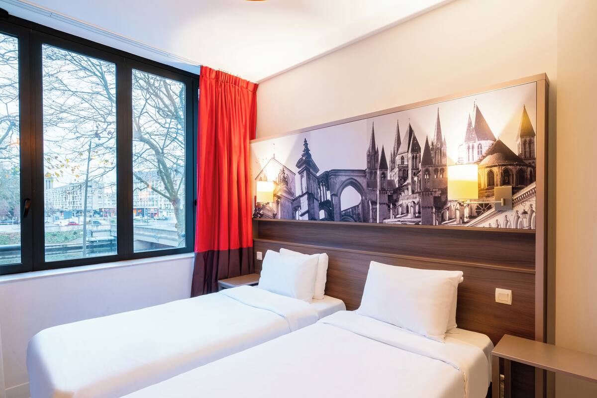 Quarto de hotel com duas camas, uma janela ampla com vista para a paisagem urbana e um quadro pendurado na parede retratando um edifício histórico.