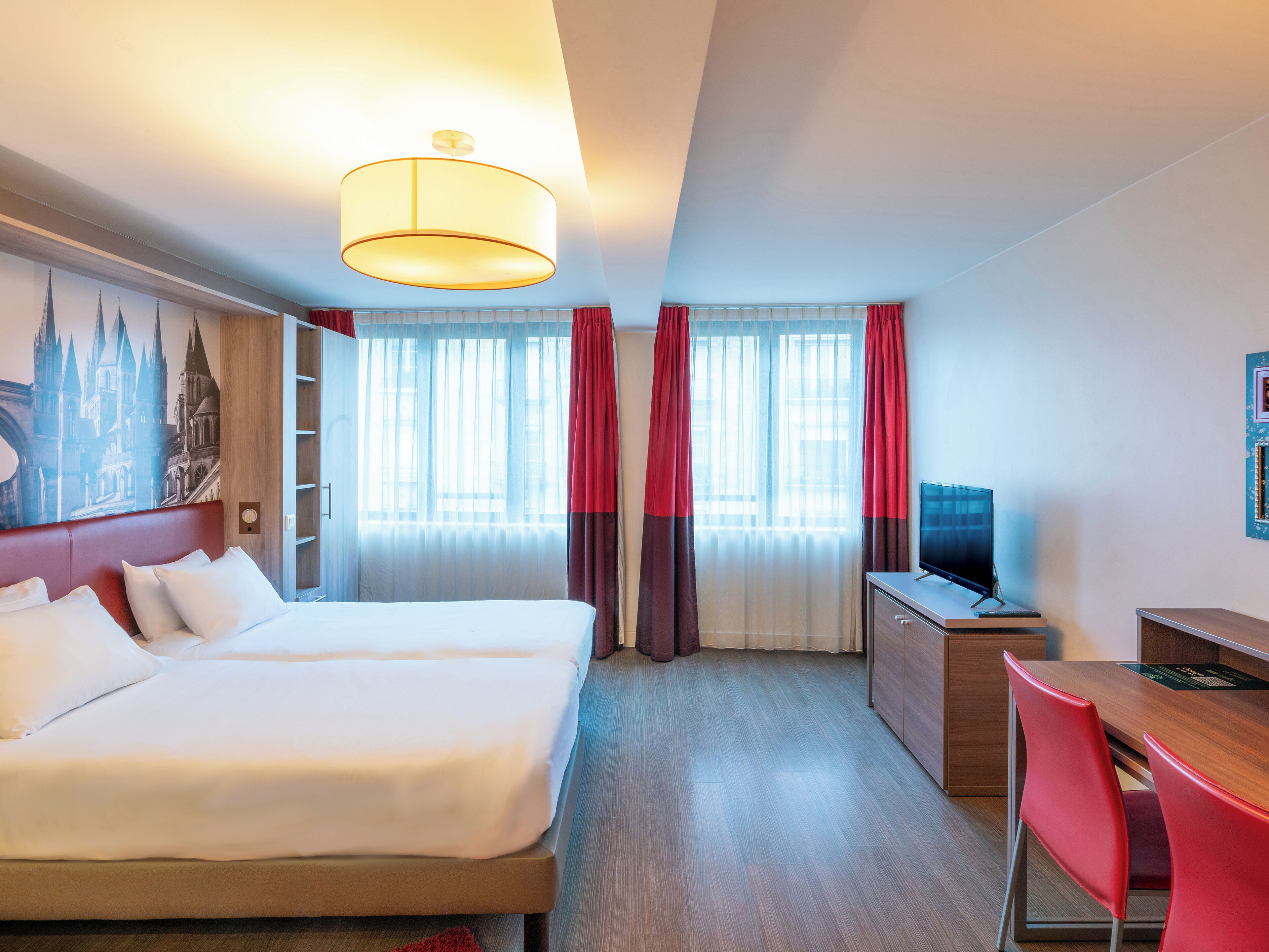 Habitación de hotel con cama, escritorio, televisor y un amplio ventanal con cortinas.