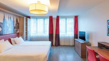 Habitación de hotel con cama, escritorio, televisor y un amplio ventanal con cortinas.