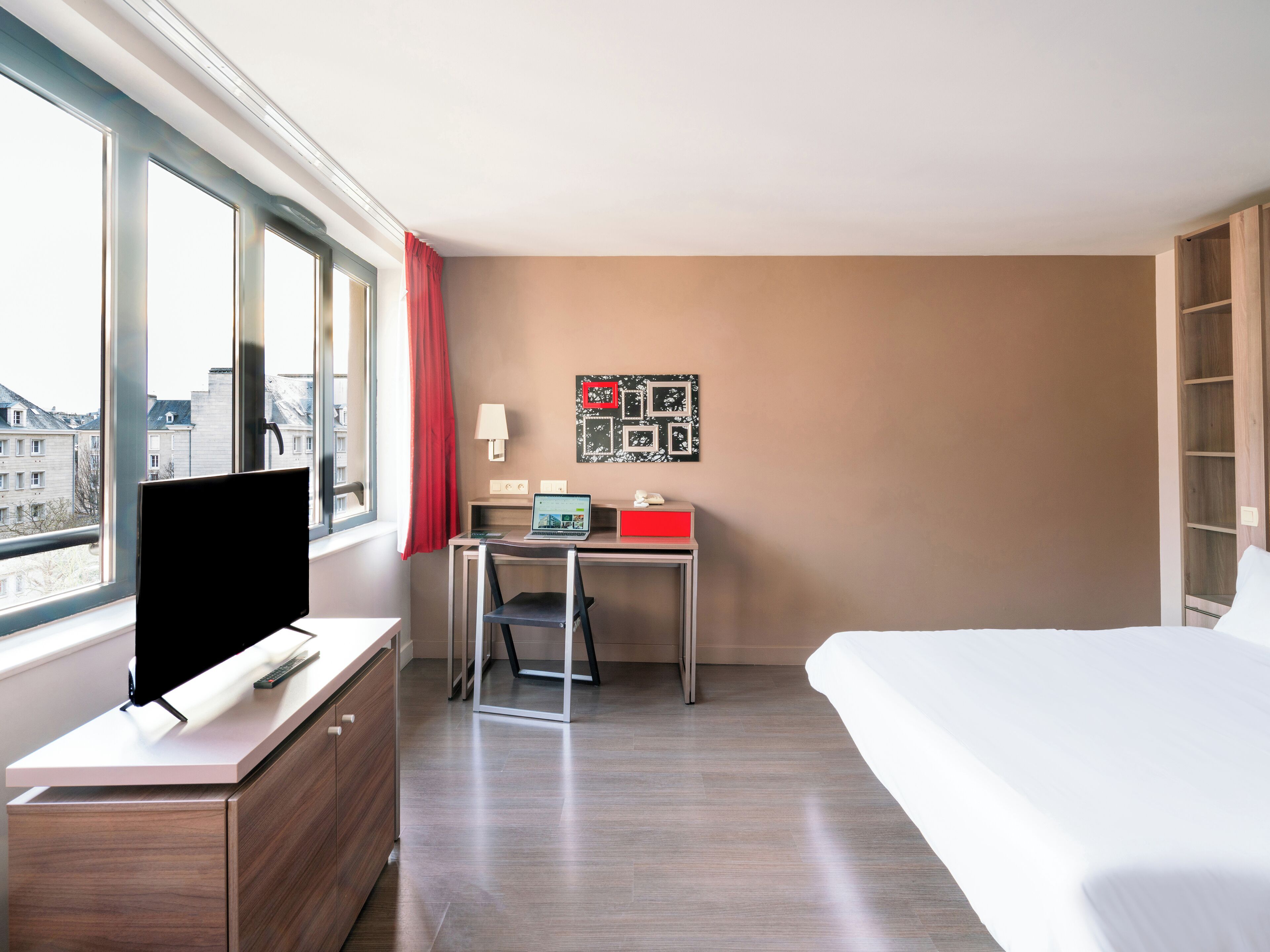 Habitación de hotel con cama, escritorio, computadora portátil, televisión y ventana con vista a edificios.