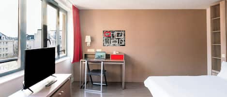 Habitación de hotel con cama, escritorio, computadora portátil, televisión y ventana con vista a edificios.
