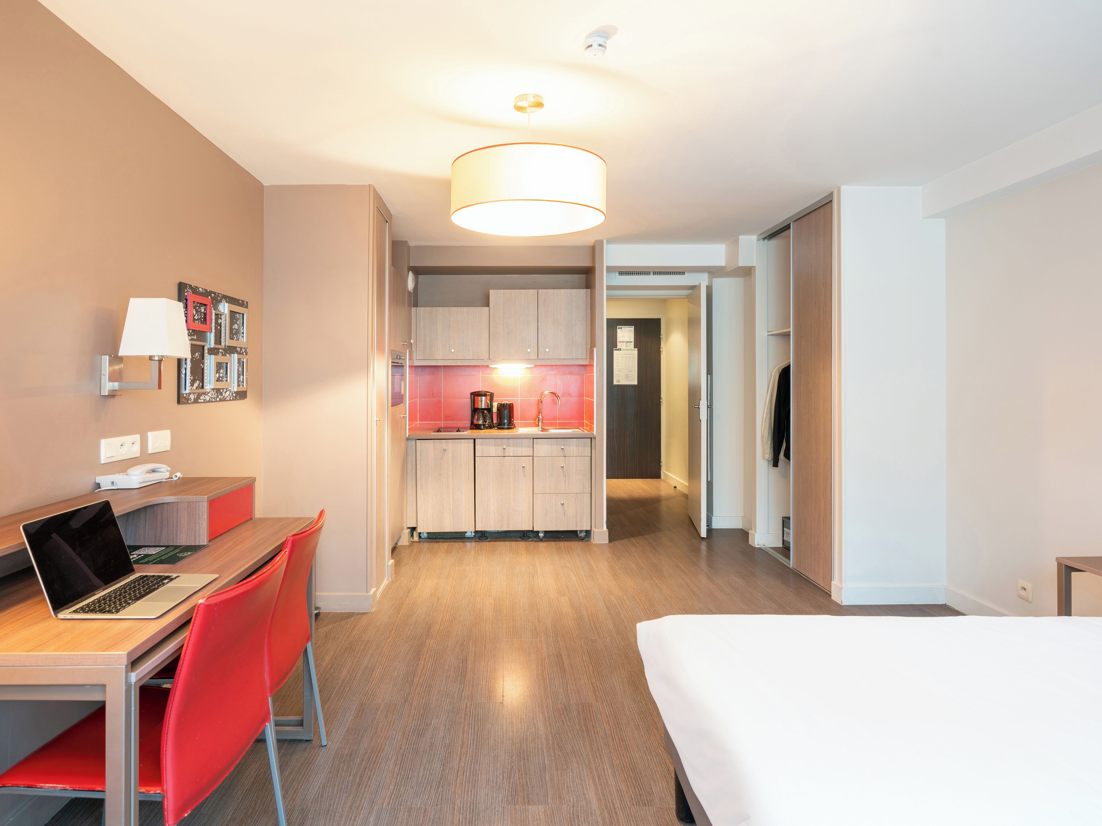 Quarto de hotel moderno com escrivaninha de madeira, uma cadeira vermelha e uma cama. Possui uma cozinha compacta com cafeteira e armários.