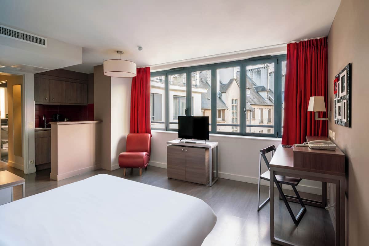 Quarto de hotel com uma cama grande, uma mesa com cadeira, televisão e vista para edifícios pela janela.