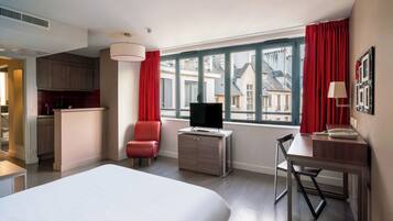 Ein Hotelzimmer mit einem großen Bett, einem Schreibtisch mit Stuhl, einem Fernseher und Blick auf Gebäude durch das Fenster.