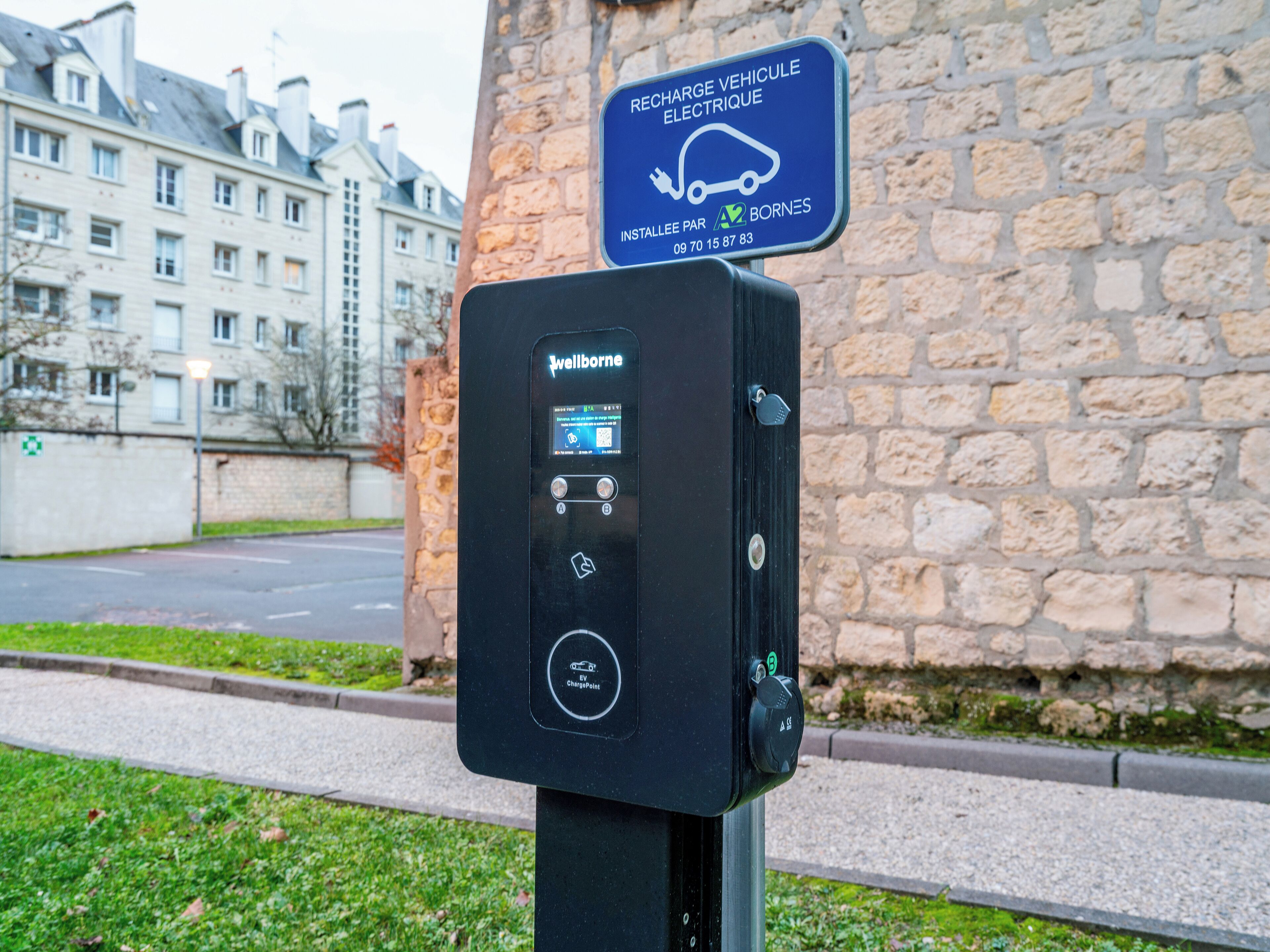 An electric vehicle charging station with a digital display, a charging point symbol, and a nearby sign indicating it is a public charging point.