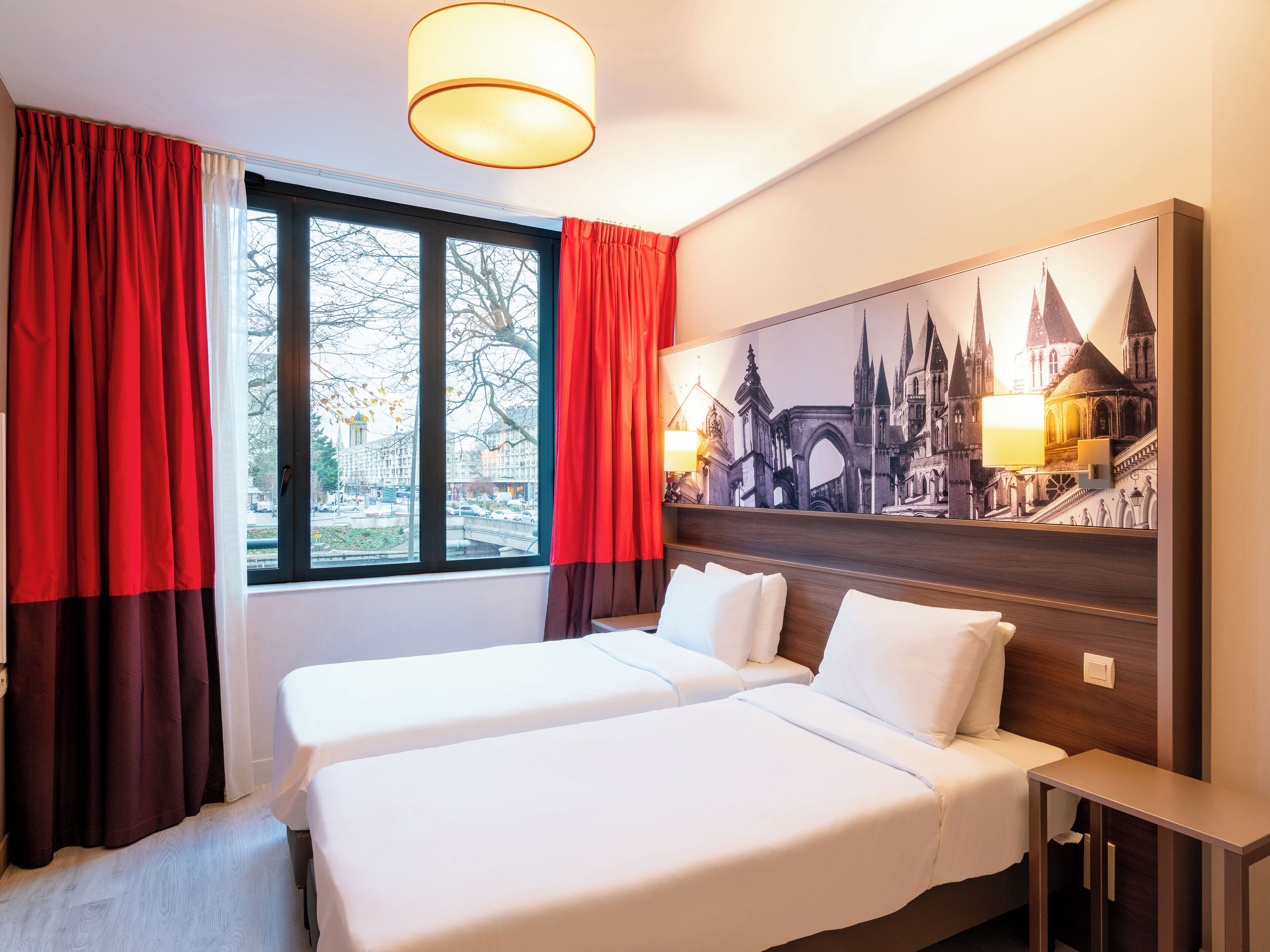Habitación de hotel con dos camas, un ventanal grande con cortinas rojas, un cuadro de un paisaje urbano en la pared y una mesita de noche con una lámpara.