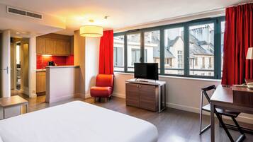 Habitación de hotel con cama, televisión, escritorio y vistas a edificios a través de amplias ventanas.