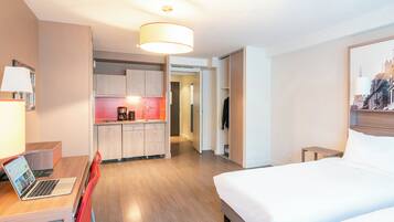 Ett modernt hotellrum med en säng, ett skrivbord med en bärbar dator, ett litet kök och en hall som leder till ett annat rum.