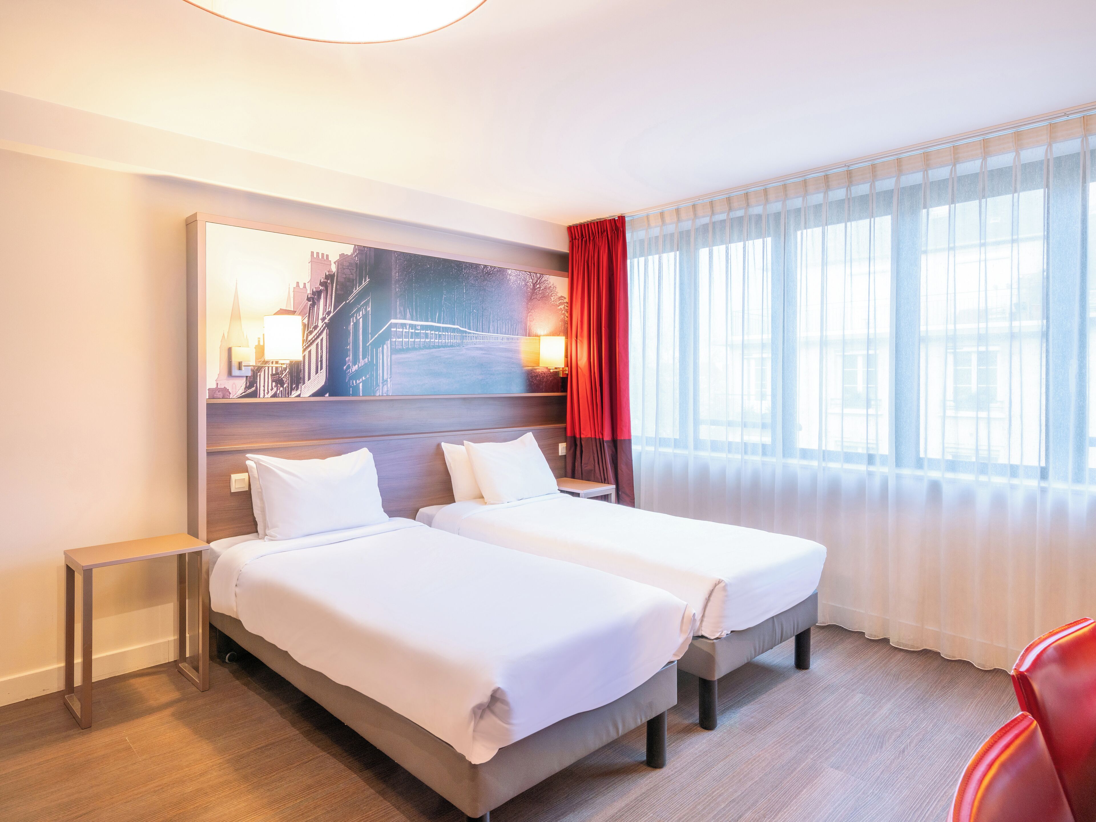 Habitación de hotel con dos camas, un ventanal con cortinas translúcidas, una silla roja y un cuadro de paisaje urbano en la pared.