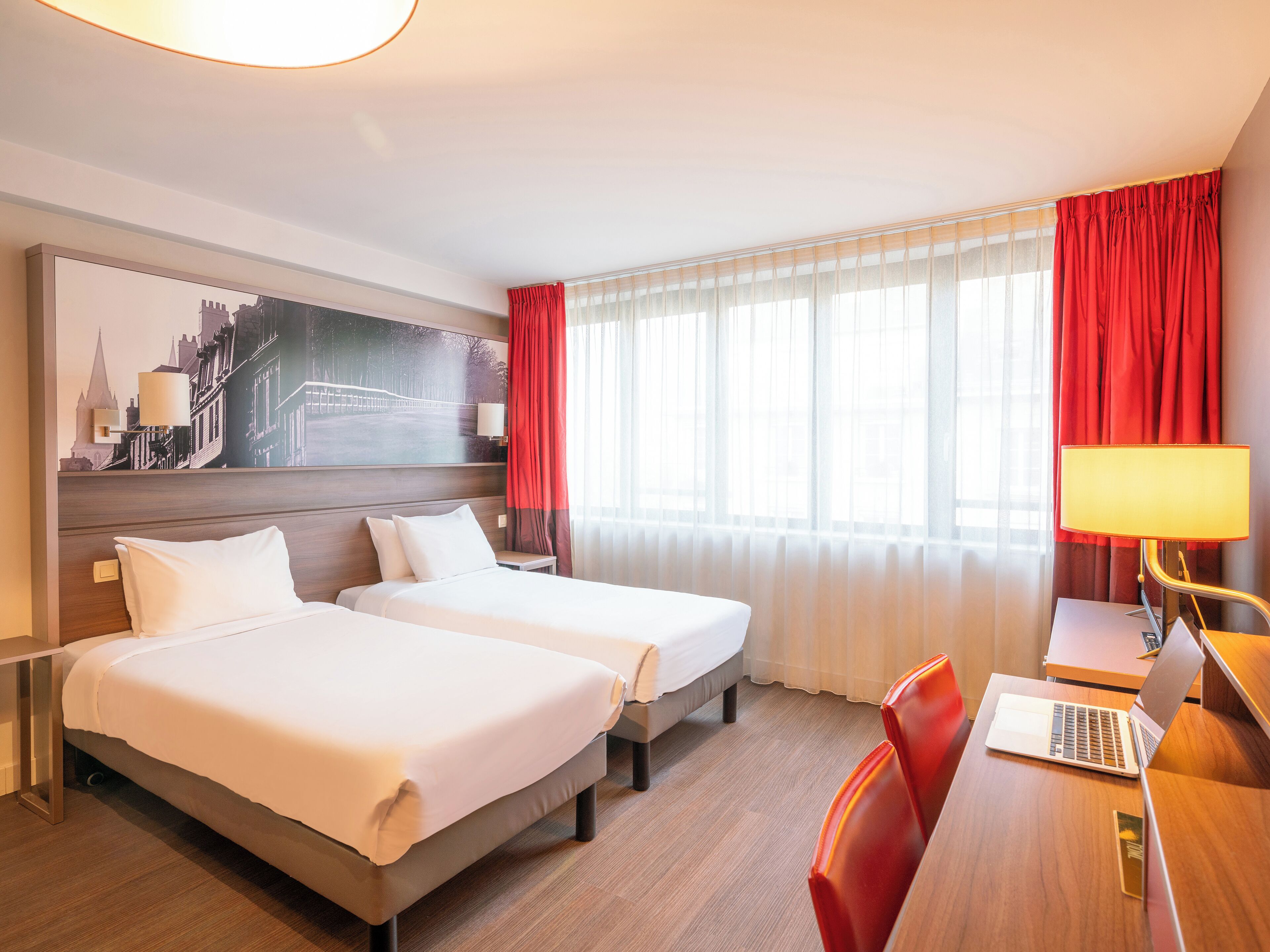 A hotel room with two beds, a desk with a laptop, a lamp, and a large cityscape painting on the wall.