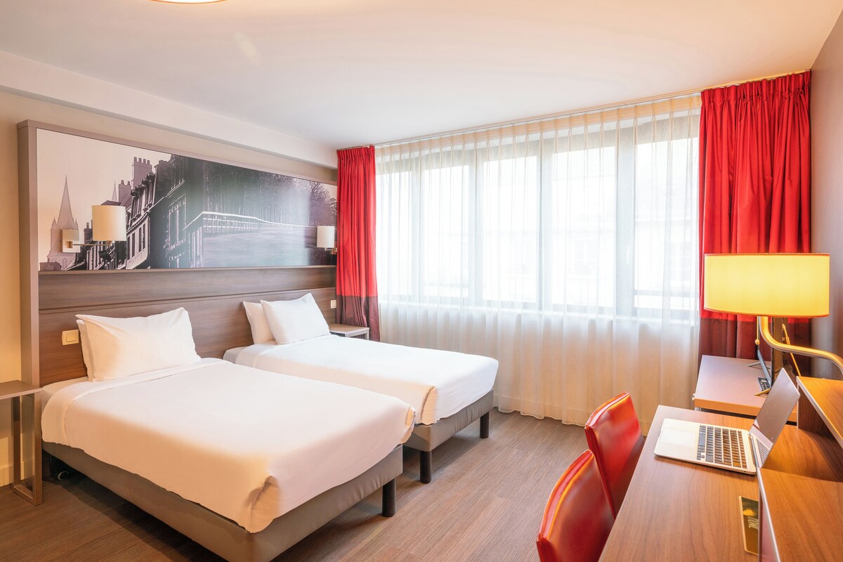 Habitación de hotel con dos camas, un escritorio con una laptop, una lámpara y un cuadro grande de un paisaje urbano en la pared.