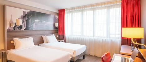A hotel room with two beds, a desk with a laptop, a lamp, and a large cityscape painting on the wall.