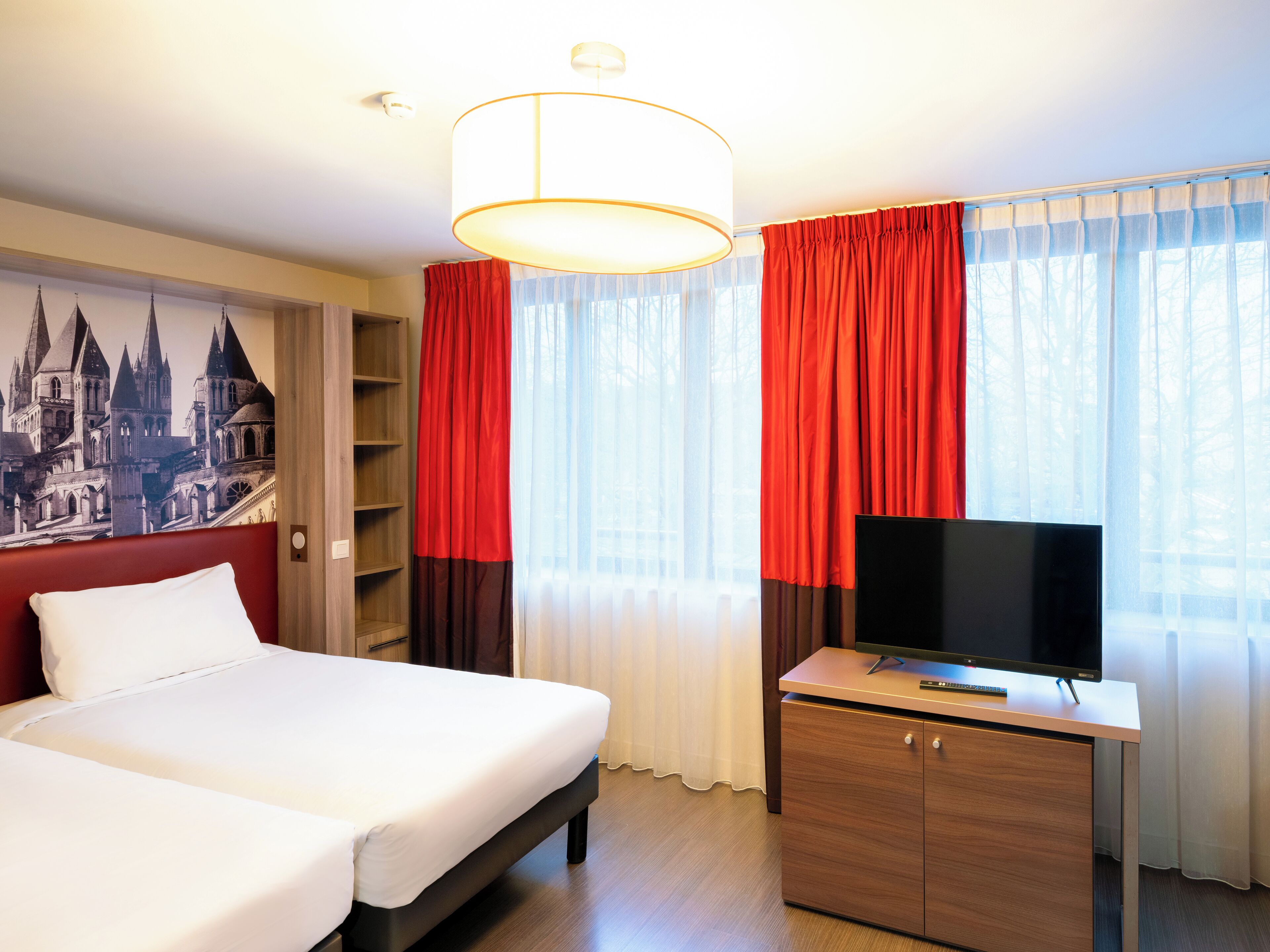 Una camera d'albergo con un letto, una TV su una scrivania in legno e un'ampia finestra con tende rosse.