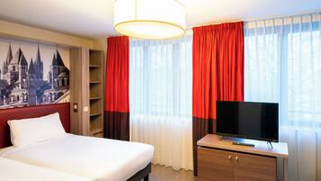 Una camera d'albergo con un letto, una TV su una scrivania in legno e un'ampia finestra con tende rosse.