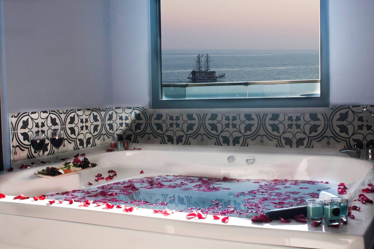 A bathtub with rose petals and glasses of drink, a window with a ship view, and patterned wall tiles.