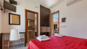 Quarto de hotel com colcha vermelha, cadeira branca, escrivaninha, televisão e guarda-roupa.
