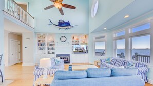 Un salon spacieux doté d’une grande fenêtre donnant sur la mer, d’un ventilateur de plafond et d’une bibliothèque remplie de livres.