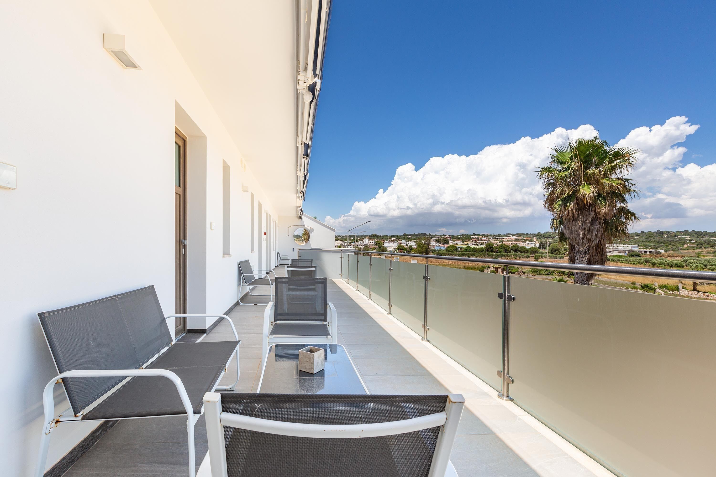Un balcón con una fila de sillas y una mesa, con vistas a un paisaje costero con una palmera y edificios a lo lejos.