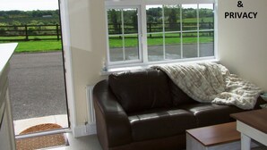 Une pièce avec un canapé en cuir marron, une couverture blanche et une vue sur un paysage verdoyant, visible à travers de grandes fenêtres.