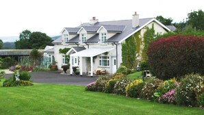 Une grande maison à deux étages, dotée d’un patio couvert, entourée de jardins bien entretenus et accessible par une allée.