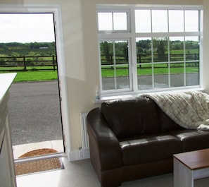 A room with a brown leather sofa, a white blanket, and a view of a green landscape through large windows. - Wonderful Setting Farmhouse Apartment (Roundwood)