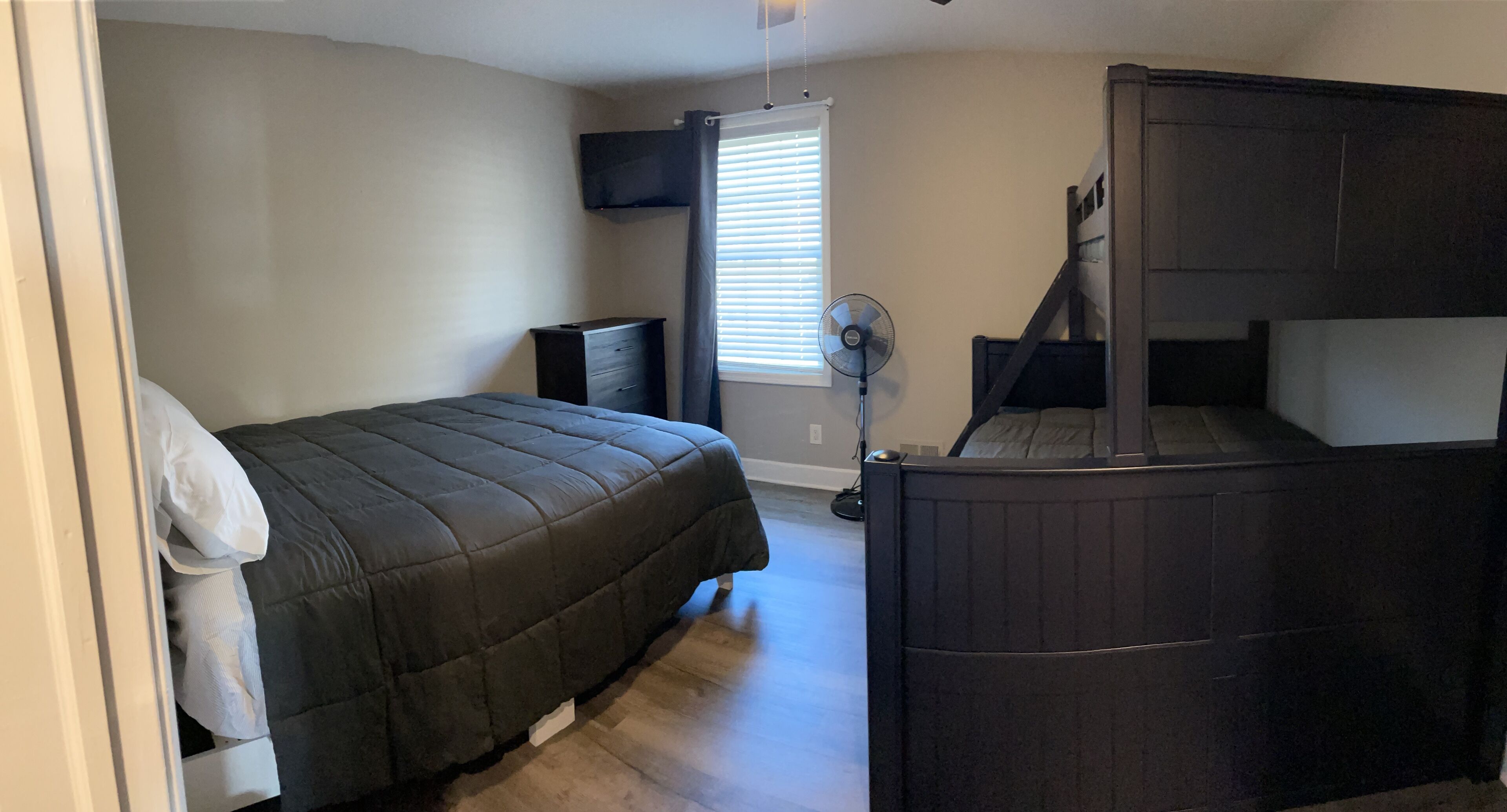 Une chambre à coucher comprenant un lit, une commode, un téléviseur fixé au mur, une fenêtre avec des stores et un ventilateur.