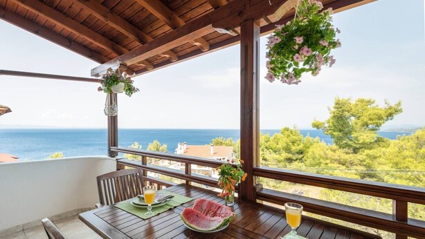 A wooden table with a view of the sea, a hanging flower arrangement, and a slice of watermelon on the table. - Beautiful and Modern Apartment in Neos Marmaras (Sithonia)