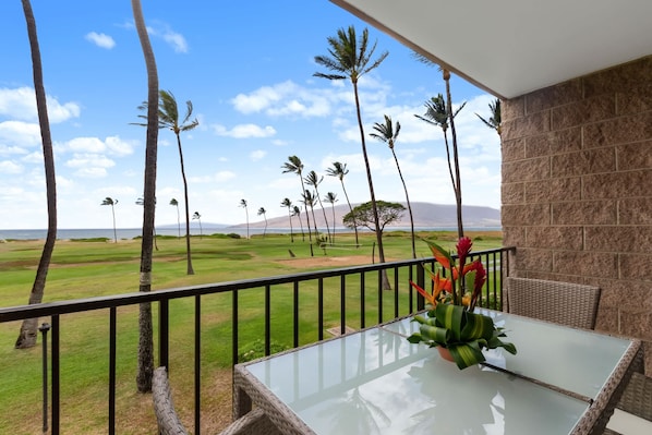 A balcony with a glass table and wicker chair, overlooking a grassy area with palm trees and a distant view of mountains.