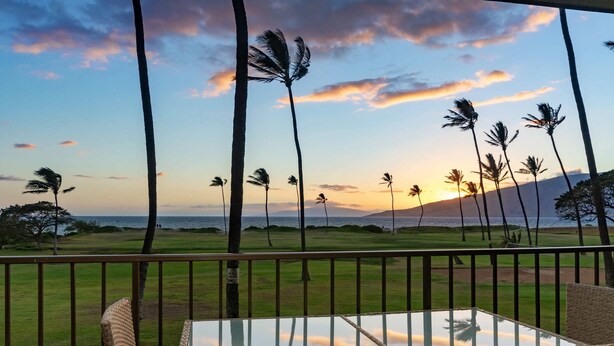 A balcony with wicker furniture, a glass table, and a view of palm trees and a sunset over a golf course.