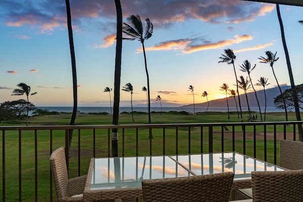 A balcony with wicker furniture, a glass table, and a view of palm trees and a sunset over a golf course.
