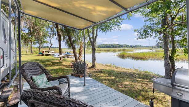 Eine überdachte Terrasse mit Korbmöbeln, einem Grill und Blick auf einen See.