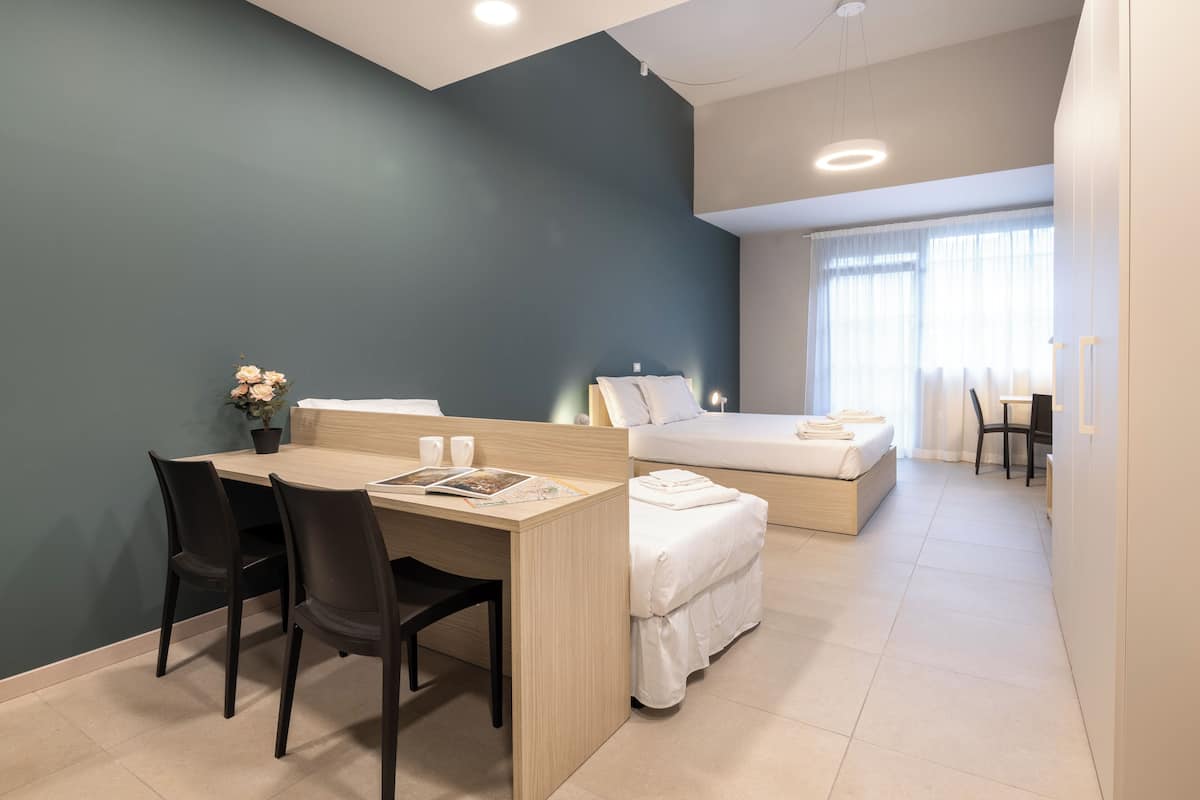 A modern hotel room with a desk, two chairs, a bed, and a small table with a plant.