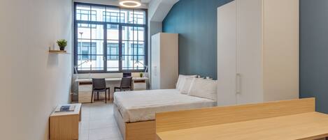 A modern bedroom with a bed, a desk, and a chair. There is a window with a view of buildings outside.
