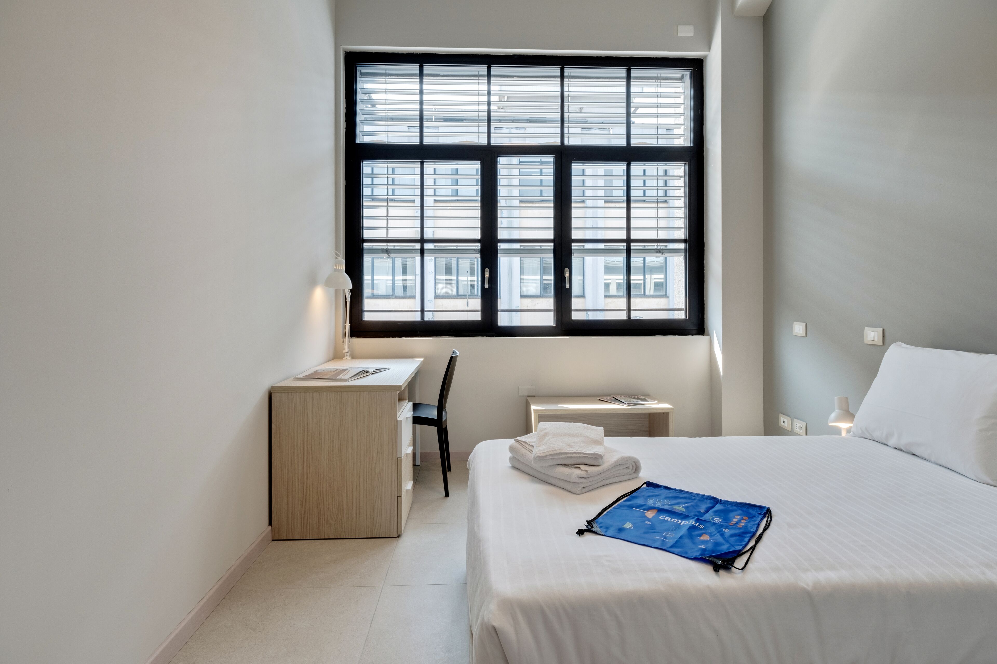 A hotel room with a bed, a desk, a chair, and a window with blinds.