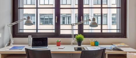 A modern workspace with a large window, two desks, chairs, a laptop, a smartphone, a plant, and various desk accessories.